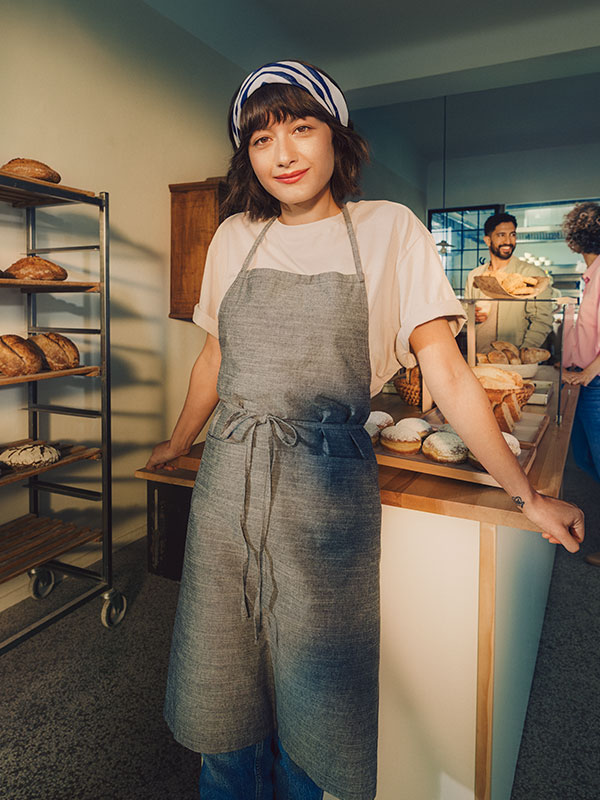 Eine Bäckerin steht in ihrer Backstube. Sie trägt eine graue Schürze und ein Kopftuch.