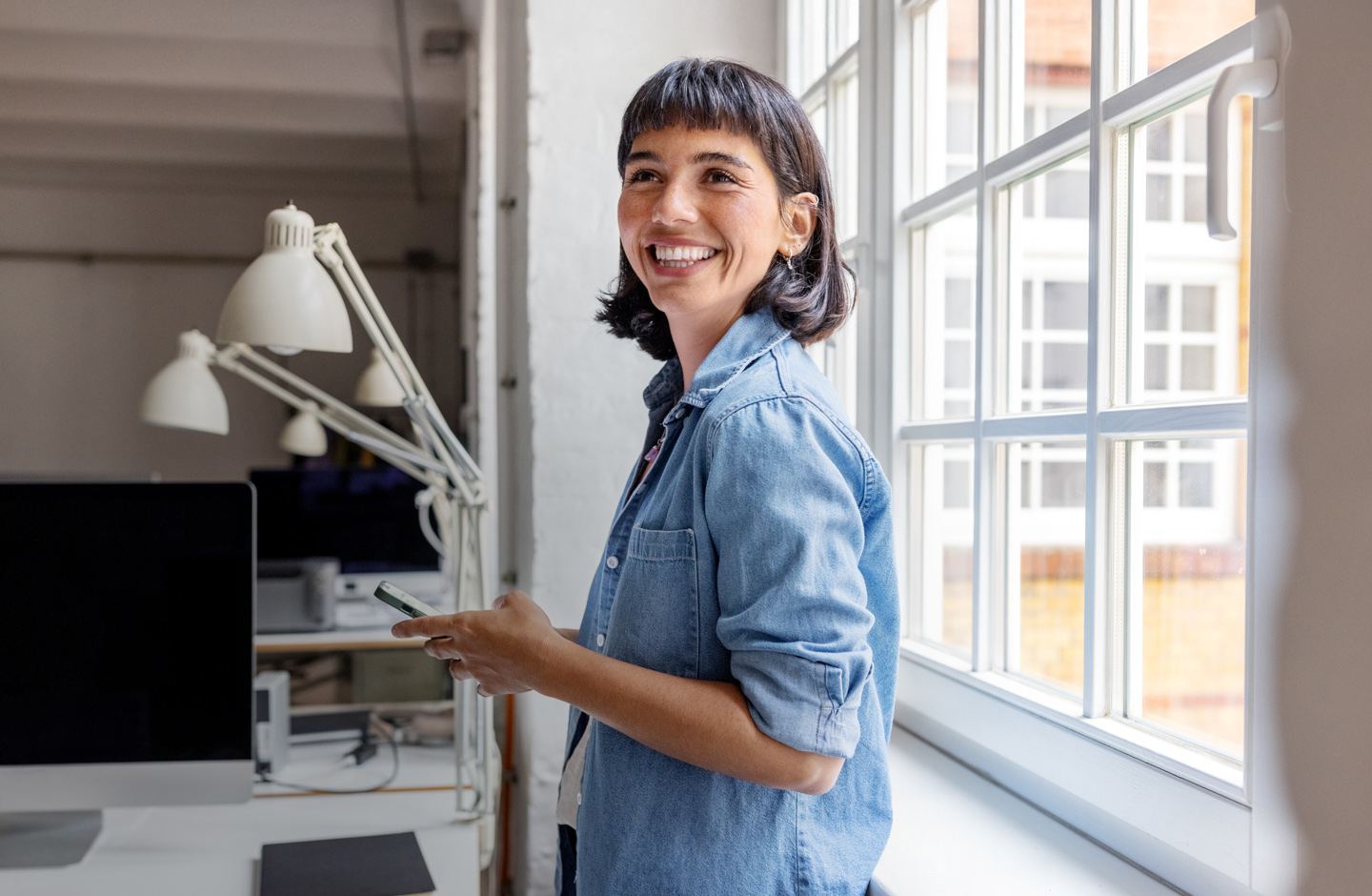 Eine junge Frau steht vor einer Fensterfront im Büro und hält lachend ihr Handy in der Hand.
