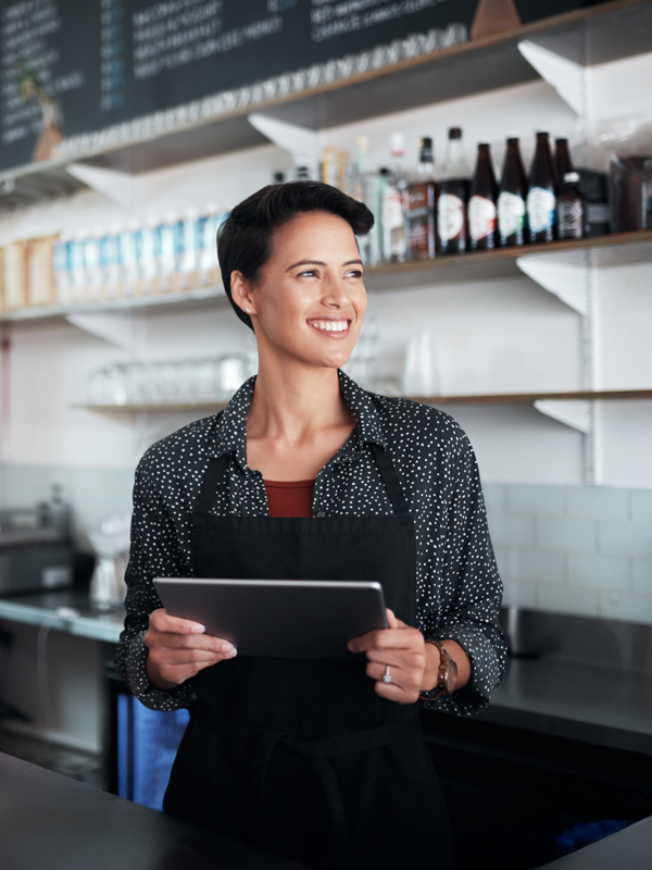 Eine junge Unternehmerin steht in ihrem Kaffeehaus. Sie lächelt und hält ein Tablet in den Händen.
