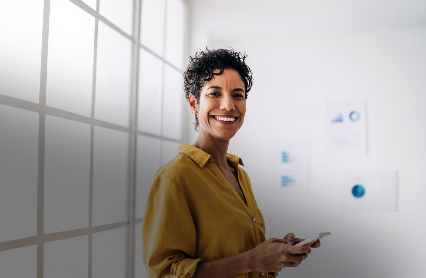 Eine Frau mit kurzem Haar und gelber Bluse steht vor einer Fensterfront mit Handy in der Hand.