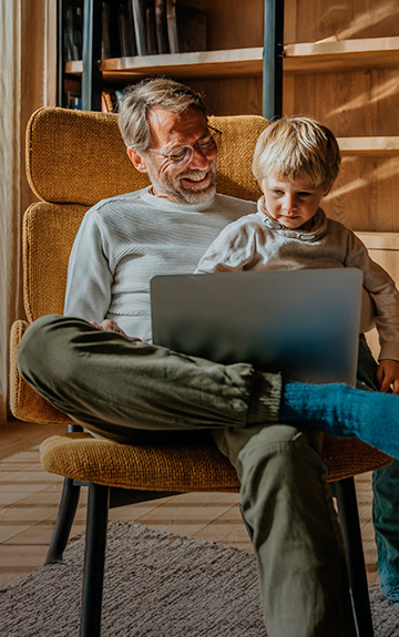 Ein Mann und ein Junge sitzen in einem Sessel und blicken gemeinsam auf ein Notebook. Der Mann lächelt, das Kind blickt konzentriert.