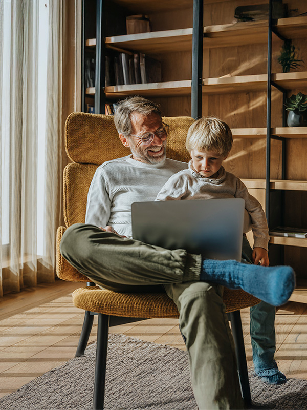 Ein Mann und ein Junge sitzen in einem Sessel und blicken gemeinsam auf ein Notebook. Der Mann lächelt, das Kind blickt konzentriert.