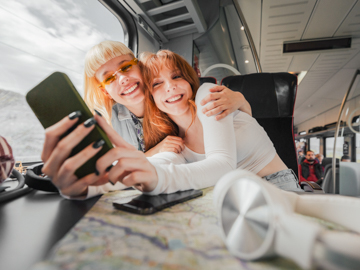 Zwei junge Frauen sitzen in einem Zug. Sie umarmen sich und blicken gemeinsam auf ein Mobiltelefon.