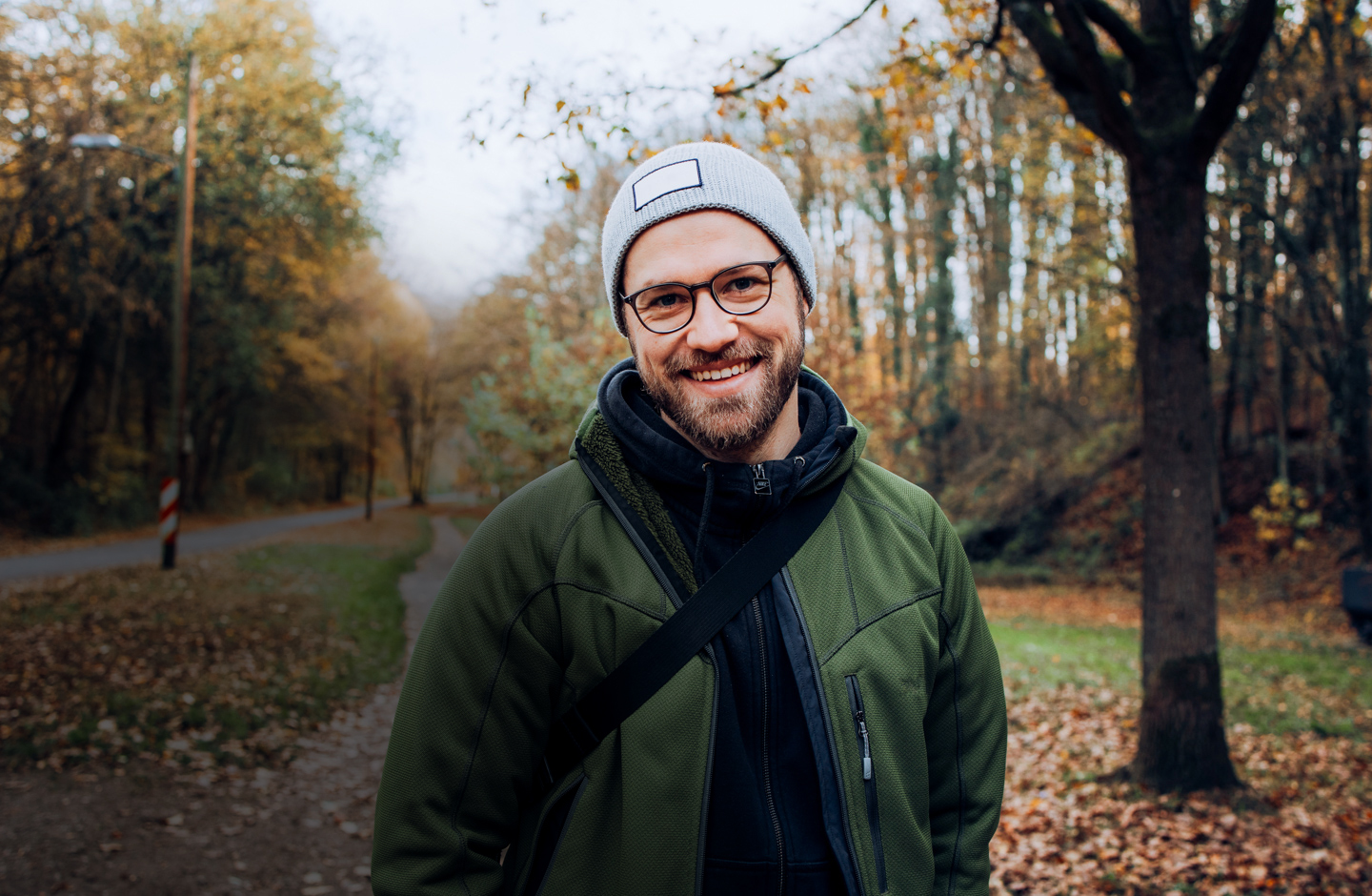 Ein Mann mit Mütze und Daunenjacke steht auf einem Spazierweg im Park und grinst in die Kamera.