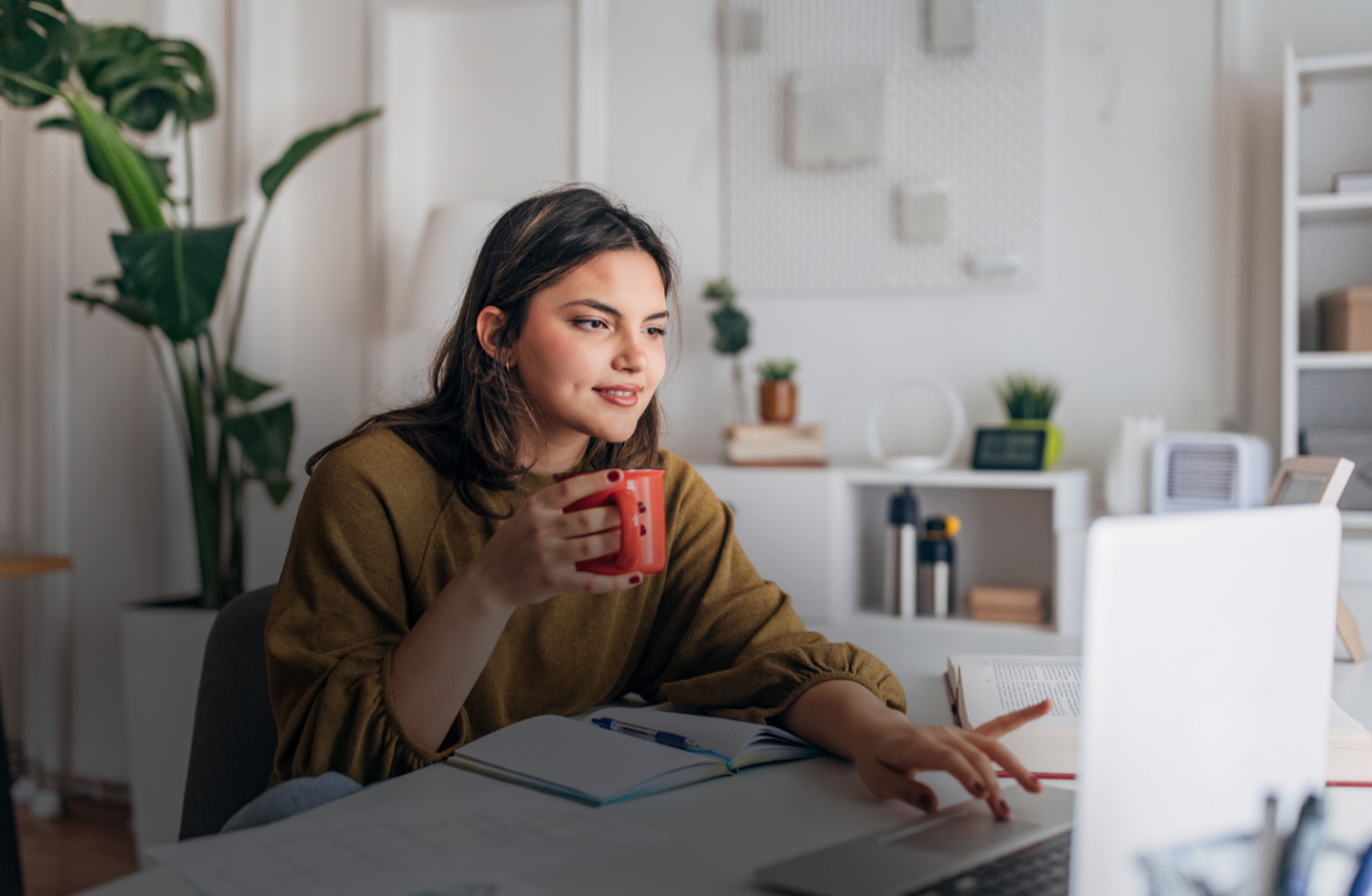 Eine junge Frau tippt mit Kaffeetasse in der Hand auf ihrem Laptop herum.